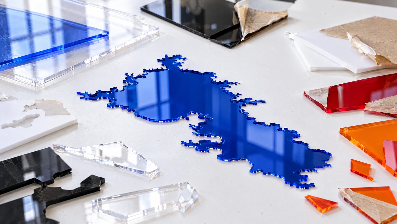Acrylic sheets and laser-cut offcuts in clear, blue, black, white, red and orange, with a glossy blue lake shape in the middle